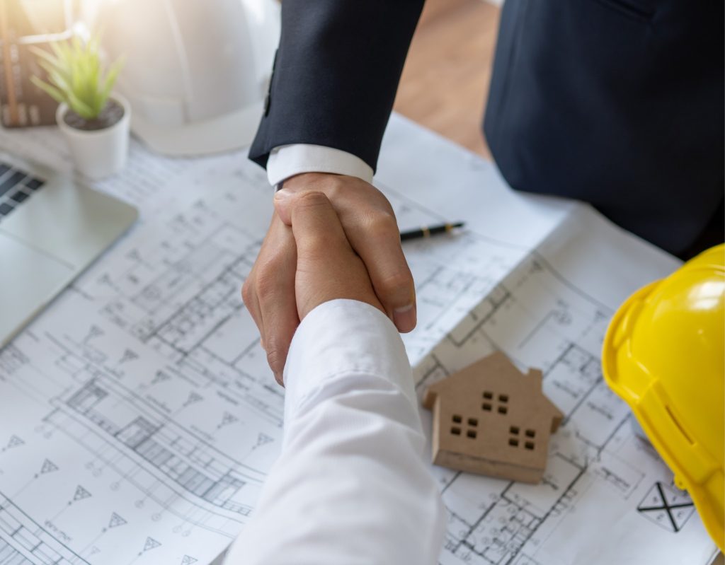 2 hommes qui se serrent la main, au dessus d'un bureau avec des plans de construction, un casque et des outils de travail