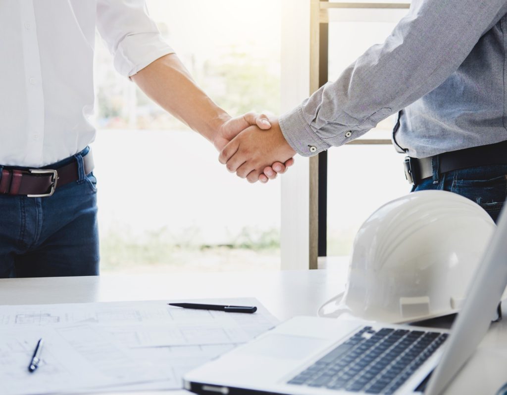 Deux hommes qui se serrent la main devant un bureau avec PC portable, plans de construction